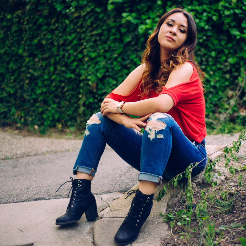Girl in blue pants and red shirt wearing boots and sitting on a cement curb
