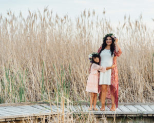 Mother and daughter wearing dresses with flowers in their hair standing on a wood platform with tall grass behind them