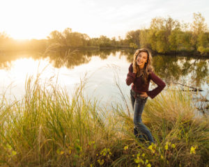 Girl wearing a purple shirt standing next to a lake with trees and a sunset in the background