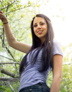 Girl wearing a blue shirt with black hair looking down at the camera with trees in the background