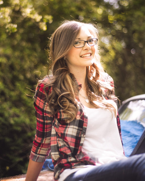 Portrait, girl in a red plaid shirt wearing glasses and sitting with green trees in the background