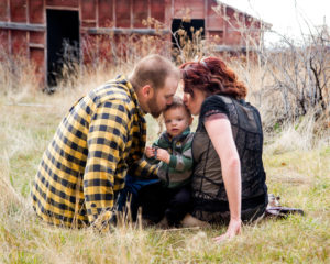 Picture of Mom and Dad kissing the top of their sons head with a red barn in the background