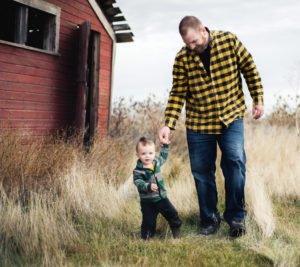 Portrait, Moss Image, Moab Photographer, Dad in a plaid yellow shirt holding a little boys hand wearing a striped green jacked with a barn in the background.