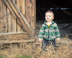 picture of a toddler in a striped green jacket standing in front of a barn.